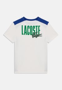 T-shirt blanc avec des accents bleus aux épaules. Présente le grand texte "LACOSTE" en vert et un logo crocodile brodé dans le dos. Matériau en coton.