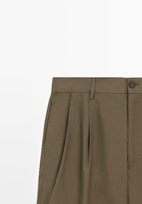 Pantaloni plissettati verde oliva con tasche anteriori, passanti per cintura, chiusura con bottone e zip su sfondo bianco.