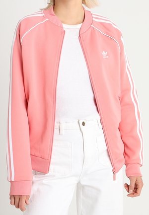 Rosa träningsjacka med vita ränder längs axlarna och ärmarna. Har dragkedja framtill, ribbade ärmslut och en broderad logotyp på bröstet.