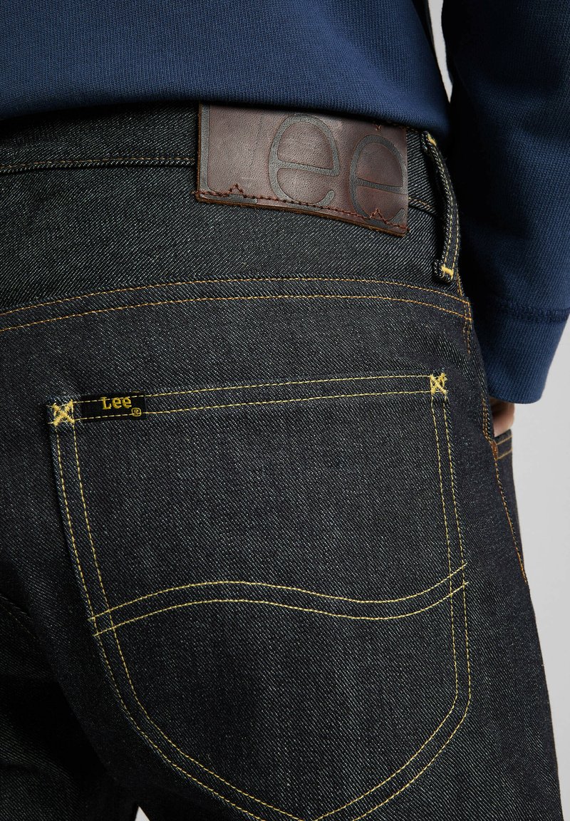 Jeans de mezclilla oscura con costuras amarillas, que cuentan con un parche de cuero marrón en la cintura embossada con "Lee" y una pequeña etiqueta negra en el bolsillo trasero.