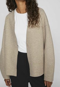 Beige cardigan i mjukt tyg, med avslappnad passform, öppen front och två knappar. Bärs över en vit skjorta och svarta byxor.