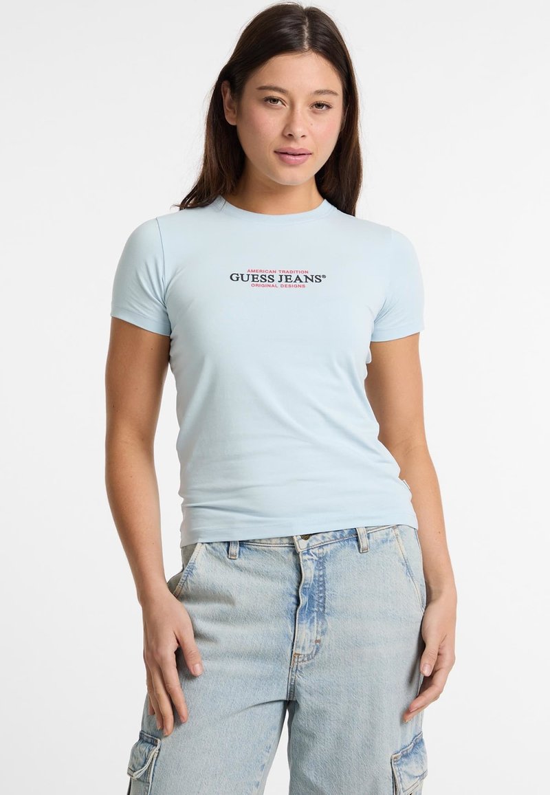 Camiseta ajustada de color azul claro con mangas cortas, que presenta un logotipo gráfico centrado en texto rojo y negro. Combina con unos vaqueros de tiro alto en un lavado claro.