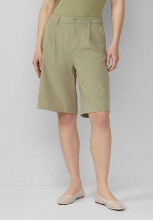 Person iført knælange beige shorts, en lysegrøn top og beige teksturerede ballerinaer, stående mod en ensfarvet grå baggrund.