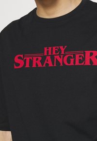 Černé bavlněné tričko s výrazným červeným nápisem "HEY STRANGER" umístěným uprostřed, s uvolněným kulatým výstřihem.