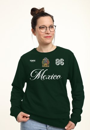 Kvinde med briller iført en mørkegrøn sweatshirt med "Mexico," et fodboldemblem, nummer 86 og "TOFFS" logo, som holder i sweatshirtens kant.