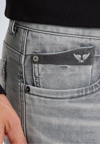 Jeans de mezclilla gris con un bolsillo frontal que muestra una etiqueta de tela gris oscuro con alas blancas y el nombre de la marca. Tejido texturizado con un aspecto desgastado.