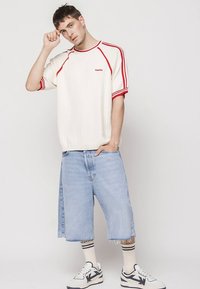 Biała prążkowana koszulka z czerwonymi akcentami, zestawiona z jasnoniebieskimi krótkimi spodniami dżinsowymi i białymi trampkami z czarnymi detalami. Modelka pozuje pewnie.