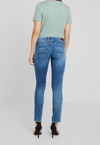 Jeans skinny di denim blu con vita media, due tasche posteriori e una toppa in pelle marrone sul retro. Abbinati a una t-shirt verde e tacchi neri.