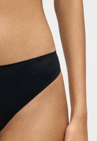 Gros plan du bas du torse d'une personne portant un bas de bikini noir uni sur un fond clair uni.