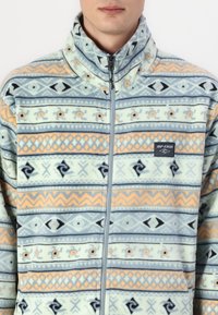 Veste en polaire à col haut, avec un motif géométrique bleu et orange, fermeture éclair à l'avant et logo de la marque sur la poitrine gauche. Texture douce.