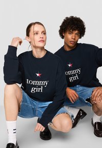 Marine sweatshirt med "tommy girl" tekst, stjerne detalje. Båret med slidte denim shorts. Modellerne har lys og mørk hår; stående og knælende poser.