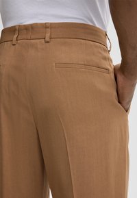 Close-up of a person wearing brown trousers with a back pocket, one hand in the side pocket, and a white shirt tucked in.