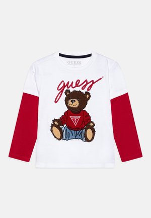 Biała koszula z długim rękawem i czerwonymi rękawami, z siedzącym misiem w czerwonym swetrze Guess i niebieskich dżinsach, z napisem „guess” nad nim.