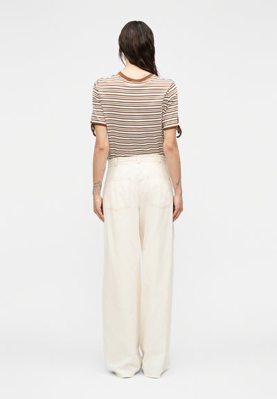Γυναίκα με μακριά σκούρα μαλλιά που φοράει κοντομάνικη μπλούζα με καφέ και λευκές ρίγες και ψηλόμεσο φαρδύ παντελόνι εκρού, φωτογραφημένη από πίσω.