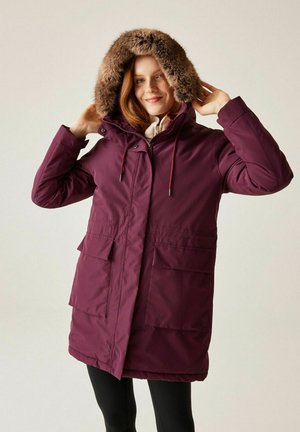 Kobieta poprawiająca kaptur długiego fioletowego zimowego płaszcza z futrzanym obszyciem, ubrana w czarne legginsy na jednolitym tle.