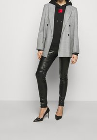 Blazer de cuadros gris de doble botonadura sobre una sudadera negra con un logo rojo, combinado con pantalones de cuero ajustados negros y tacones de aguja negros.