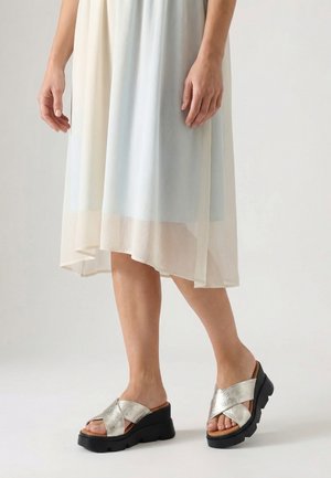 Benen met zilverkleurige plateauzandalen met gekruiste bandjes en een lichte, vloeiende midi-jurk met een asymmetrische zoom tegen een effen achtergrond.