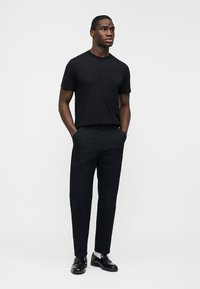 Mann steht mit den Händen in den Taschen, trägt ein schwarzes T-Shirt, schwarze Hose, weiße Socken und schwarze Lederschuhe vor weißem Hintergrund.