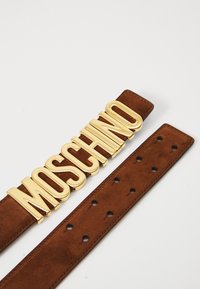 Bruine suede riem met een grote goudkleurige logo-buckle met de tekst "MOSCHINO." De riem heeft gelijkmatig verdeelde gaten voor aanpassing.