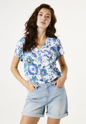 Kurzarmpullover mit V-Ausschnitt in einem blauen und grünen Blumenmuster auf weißem Hintergrund, kombiniert mit hellblauen Shorts mit umgeschlagenem Saum.