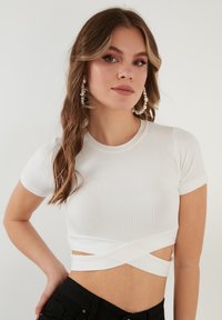Valge ribadega lühikeste varrukatega crop top, millel on seotud eesmine disain. Omaduseks on tihe lõige ja sile tekstuur.