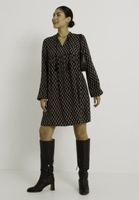 Vestido negro de manga larga con un diseño estampado, que presenta pliegues y un escote en V. Combinado con botas de cuero marrón de tacón alto hasta la rodilla.