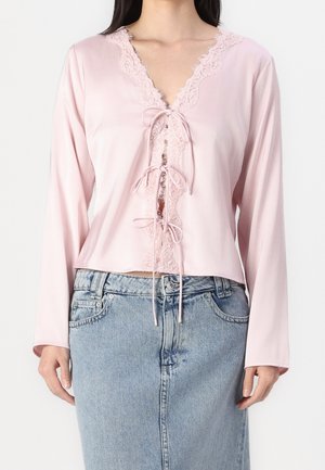 Vrouw draagt een lichtroze blouse met lange mouwen, kanten rand en strikjes aan de voorkant, gecombineerd met een hooggetailleerde lichtblauwe spijkerrok.