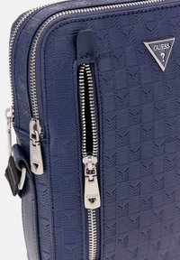 Marinblå texturerad zippered väska med fiskbensmönster, med silverfärgade metallaccenter och en triangel-logotagg. Slät syntetisk material.