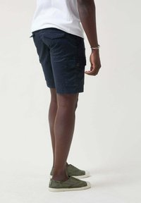 Pantalones cortos cargo azul marino con un corte relajado, que cuentan con bolsillos laterales y un bolsillo trasero, hechos de una tela ligera y duradera. Llevados con zapatos verde tipo slip-on.