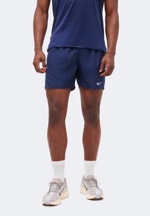 Mężczyzna w granatowych sportowych szortach, pasującej koszulce, białych skarpetkach i szarych butach do biegania stoi na jednolitym tle.