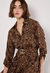 Robe imprimée léopard dans des nuances de marron, avec un col à revers et des manches retroussées. Ceinture en double anneau doré en accent à la taille.