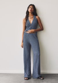 Grau gemusterter Jumpsuit mit tiefem V-Ausschnitt und weit geschnittenem Bein. Verfügt über ein geripptes Muster, minimales Zubehör und eine elegante Silhouette.