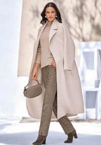 Beige lange jas met brede revers, gelaagd over een lichtbeige trui. Grijze plaid broek en een bijpassende handtas. Bruine enkellaarsjes met hak.