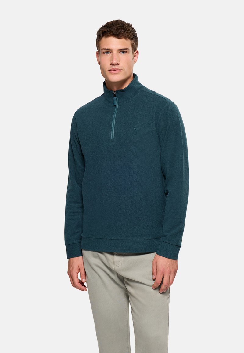 Hombre con un pulóver de cremallera hasta el pecho de color verde azulado y pantalones beige, de pie frente a un fondo liso de color gris claro.