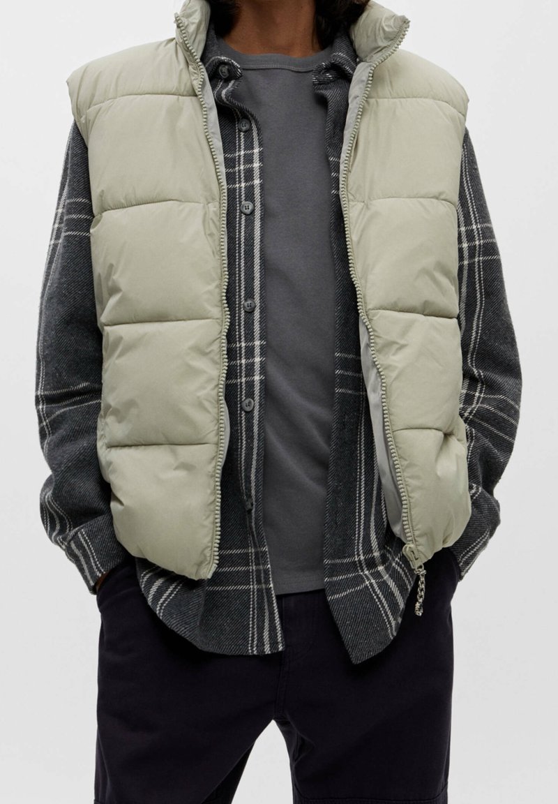 Personne portant une veste matelassée beige clair par-dessus une chemise à carreaux gris foncé déboutonnée et un t-shirt gris avec un pantalon foncé.