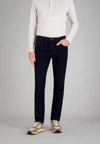 Dunkelblaue Jeans mit Slim Fit, aus Denim gefertigt. Verfügt über orangefarbene Nähte und klassische Taschen. Getragen mit einem hellgrauen Langarmshirt.