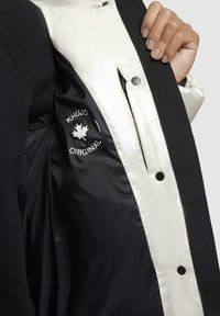 Giacca nera con un interno in satin bianco. Presenta un logo visibile sulla fodera, bottoni a pressione neri e una texture liscia.