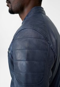 Marineblaue Lederjacke mit einem gesteppten Schulterdesign und Reißverschlusstasche. Glatte Textur mit dezenter Glanzwirkung. Figurbetonter Schnitt.