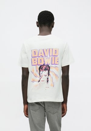 Homme de dos portant un t-shirt blanc avec un texte coloré et un portrait de David Bowie, associé à un jean gris, sur fond uni.