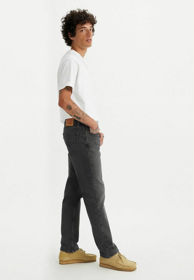 T-shirt blanc à manches courtes, jean gris foncé à jambes droites et mocassins couleur fauve. Tatouages remarquables visibles sur les bras. Tenue simple et décontractée.