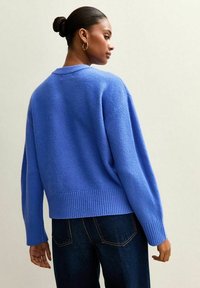 Suéter azul holgado hecho de tela de punto texturizada con dobladillo y mangas acanaladas. Se lleva con jeans de mezclilla oscura, mostrado desde atrás.