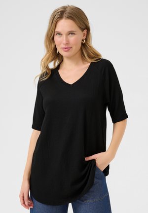 Haut noir à col en V avec des manches courtes, fabriqué en tissu texturé. Le design présente une coupe décontractée et un ourlet arrondi, souligné par une main dans la poche.