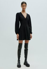 Schwarzes Kleid mit V-Ausschnitt, Knopfleiste, langen Puffärmeln und einem ausgestellten Rock. Kombiniert mit hohen schwarzen Lederstiefeln.