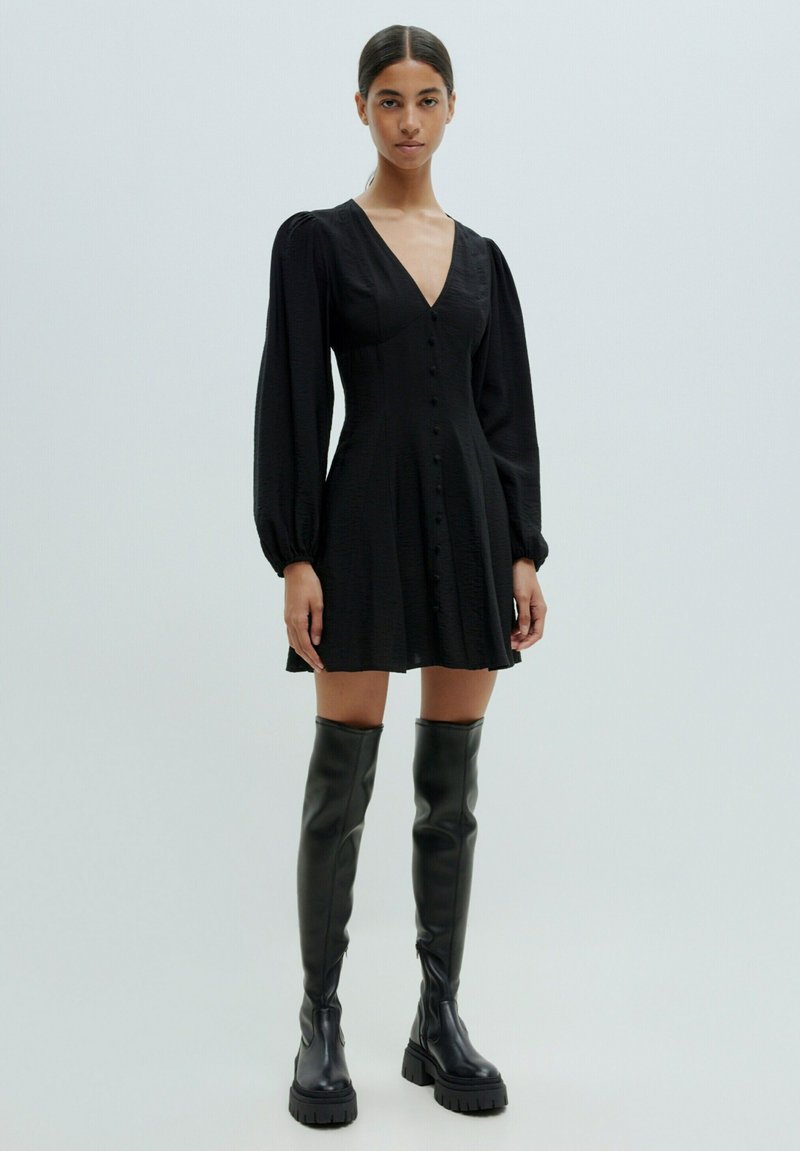 Schwarzes Kleid mit V-Ausschnitt, Knopfleiste, langen Puffärmeln und einem ausgestellten Rock. Kombiniert mit hohen schwarzen Lederstiefeln.