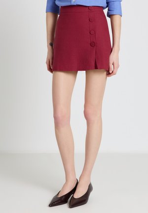 Donna che indossa una gonna corta bordeaux con bottoni laterali, abbinata a tacchi neri a punta e una camicia blu con le maniche arrotolate.