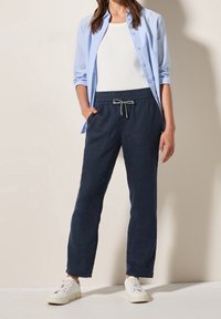 Marinblå linneblandade byxor med elastisk midja, fickor och dragsko. I kombination med en vit linne och en ljusblå skjorta med knappar, samt vita sneakers.