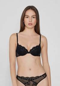 Sujetador de encaje negro con paneles transparentes y tachuelas decorativas. Presenta un borde con forma de ondas y tirantes ajustables. Combinado con un tanga de encaje a juego.
