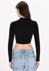 Fekete bordás, hosszú ujjú crop top, magas nyakkivágással, világoskék, magas derekú farmernadrággal párosítva. Simább textúra, testhezálló kialakítás.