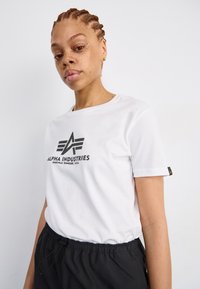 Valkoinen puuvillapaita, jossa on musta "ALPHA INDUSTRIES" -logo edessä, pyöreä kaula-aukko ja lyhyet hihat. Yhdistetty mustiin nyöritettävään housuihin.