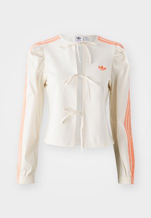 Kermainen pitkähihainen takki, jossa on etutiet, joustavat hihansuut ja oranssit kolmiraidat hihassa. Takissa on brodeerattu logo rinnassa.